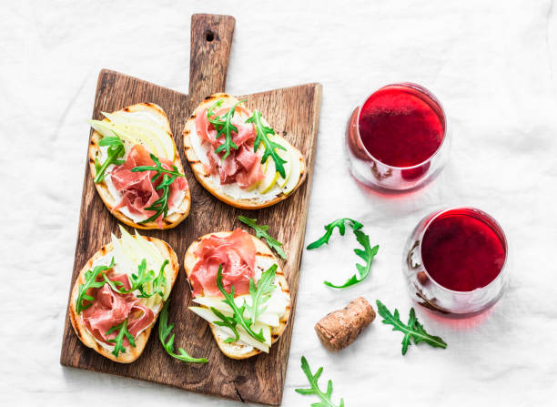 Bruschetta with cream cheese, pear, prosciutto, arugula on wooden chopping board and red wine  on light background, top view. Flat lay Bruschetta with cream cheese, pear, prosciutto, arugula on wooden chopping board and red wine  on light background, top view. Flat lay aperitif photos stock pictures, royalty-free photos & images