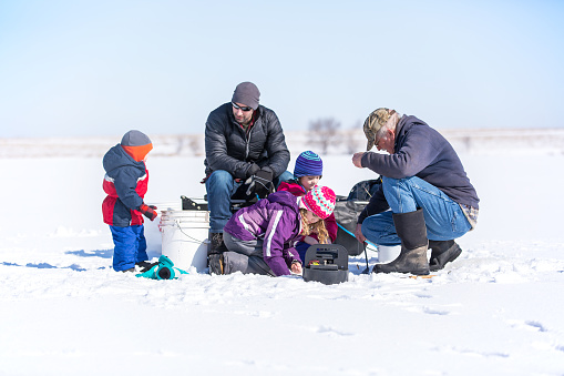 Three young children with their dad and grandpa ice fishing on a frozen lake in late winter / early springtime. The ice is covered with snow. The two sisters are kneeling near Grandpa, who is getting a fishing pole line ready for them to fish with. Dad is sitting on the portable fish house/sled, while the young boy looks on.
