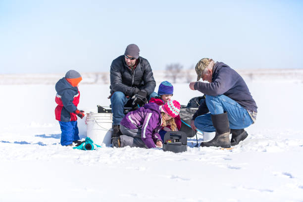 Family Ice Fishing On Late Winter Day Three young children with their dad and grandpa ice fishing on a frozen lake in late winter / early springtime. The ice is covered with snow. The two sisters are kneeling near Grandpa, who is getting a fishing pole line ready for them to fish with. Dad is sitting on the portable fish house/sled, while the young boy looks on. man ice fishing stock pictures, royalty-free photos & images