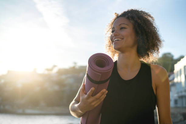 Fit woman holding a yoga mat at the beach Portrait of a fit woman holding a yoga mat at the beach and looking very happy - wellness concepts exercise mat stock pictures, royalty-free photos & images