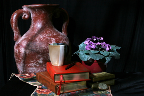 Still life to appear as light is coming from the window.  Collection of an earthern jug, leather bound books, coffee mug, aftican violets in pot, treasure box and old pocket watch on black background with a tapestry runner.