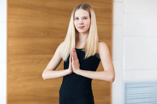 młoda blondynka kobieta w czarnym garniturze praktykujących medytację na siłowni w klasie jogi. relaks i rozciąganie. kobieta trzyma ręce w postawie namaste, patrząc na kamerę i uśmiechając się. zbliżenie - good posture yogaclass lifestyles sport zdjęcia i obrazy z banku zdjęć