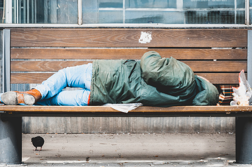 Poor homeless man or refugee sleeping on the wooden bench on the urban street in the city, social documentary concept