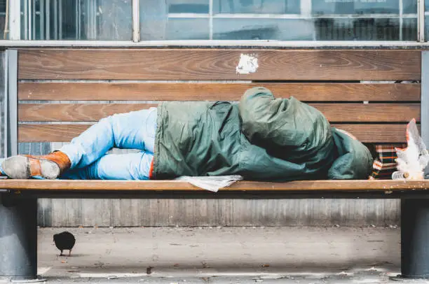 Photo of Poor homeless man or refugee sleeping on the wooden bench on the urban street in the city, social documentary concept