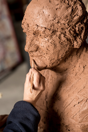 Female sculptor's hand working on the detail of a clay bust's lips. In the series, there are a variety of tools used to get different textures - palette knife, wooden block, curved spatula and straight-edged implements