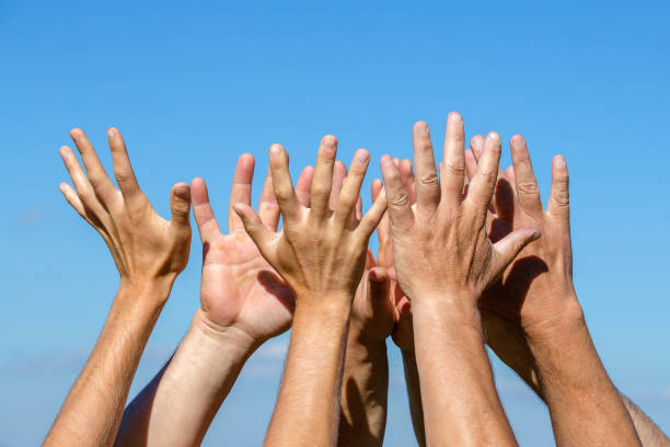 istock Group of people pulling hands in the air in sunlight. Many hands against blue sky background 940948484