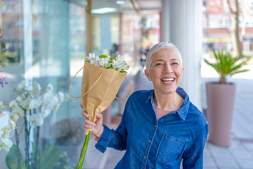 Portrait Of Happy stunning and beautiful, Mature or senior gray hair woman in denim shirt with bouquet of flowers standing outdoors during sunny day. Caucasian middle aged woman holding bunch of flowers while looking at camera with huge smile on her face. This is her day.