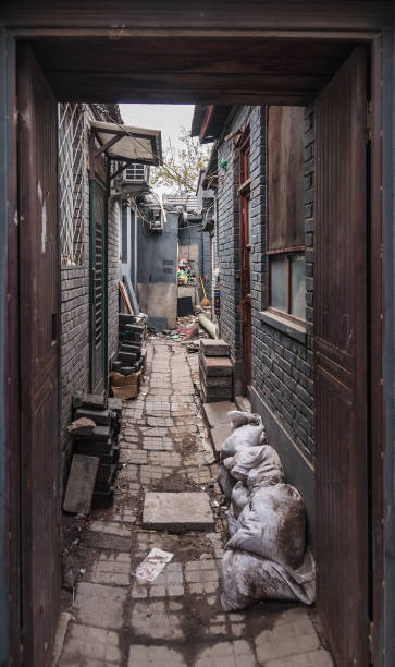 Narrow Hutong alley in Beijing. Beijing, China - April 26, 2010: Narrow Hutong alley with rubbish and uneven pavement. Dark brown wood and gray stones and dirt. View through alley entrance door. chinese slum stock pictures, royalty-free photos & images