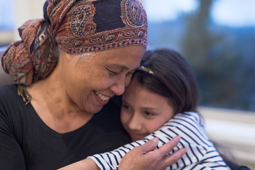 A beautiful senior woman with cancer holds her granddaughter tightly in her lap. Grandma is wearing a headscarf and smiling. Her granddaughter has a more serious expression.