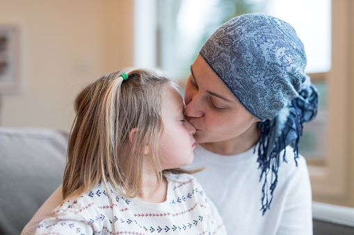 A beautiful young woman with cancer holds her preschool-age daughter in her lap by their living window. She is gently kissing her daughter's nose. Mom is wearing a headscarf.