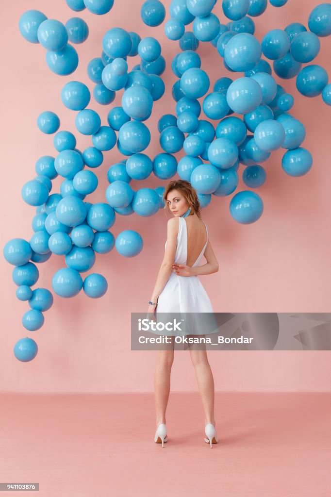 Young woman in white cocktail dress standing back to camera on pink wall background with blue bubbles hanging. Dreaming concept. Young woman in white cocktail dress standing back to camera on pink wall background with blue bubbles hanging. Dreaming concept Women Stock Photo