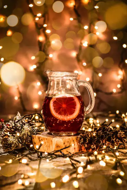 Photo of a glass carafe with fruit tea and a piece of lemon in it on a wooden table with bokeh and christmas lights all around