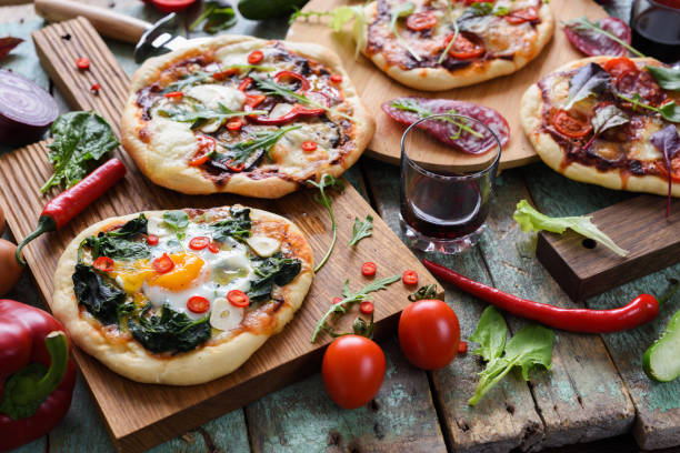 istock Pizza party. Small homemade rustic pizzas with eggs, spinach, salami, pepper, chili, arugula and tomatoes, wine glass on oak boards on shabby blue background 941117570