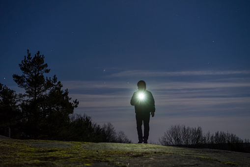 Human standing outdoor at night with flashlight and hoodie on head. Male man standing in Swedish Scandinavian nature and landscape shining with torch. Calm, peaceful photo. Mystical abstract image.