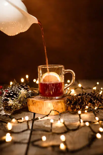 Photo of a piece of lemon in glass mug pouring tea from the tea pot with decoration, bokeh effect and christmas lights on a wooden table