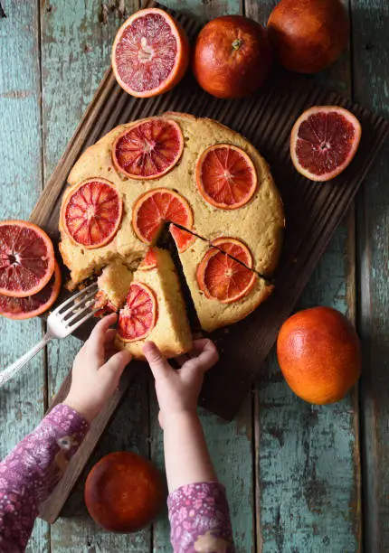 Dessert for children. Baby hands reaching for piece of blood orange cake on oak board on shabby blue background top view