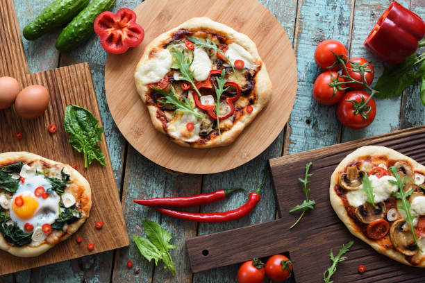 istock Pizza party flatlay. Freshly backed homemade rustic pizzas with eggs, spinach, bell pepper and mushrooms served with raw chili pepper, cucumbers and tomatoes on shabby blue background 941127890
