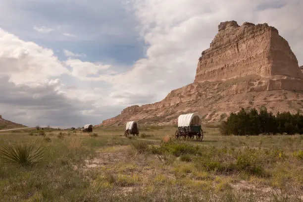 With yuccas and prairie in the foreground, clouds pass over old covered Conestoga and Murphy Wagons on the Oregon Trail with Scotts Bluff in the background in Scotts Bluff National Monument in Nebraska.