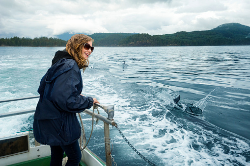 Smiling blond teenage girl with dental braces, sunglasses and rain jacket watches dolphins swimming behind the boat while cruising through the water, Pacific Ocean, Campbell River, Vancouver Island, British Columbia, Canada