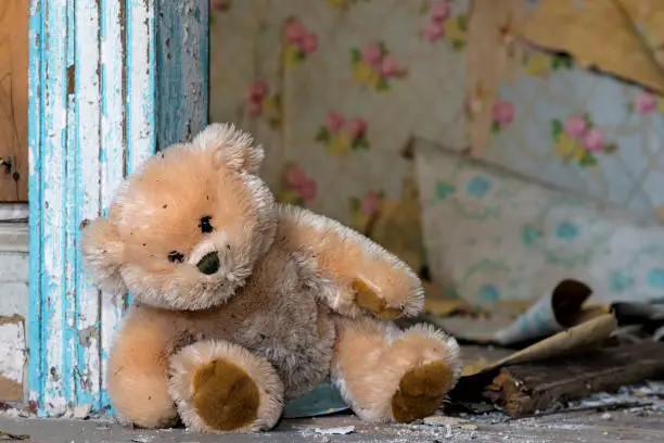 A dirty teddy bear leaning against a door frame in an old, abandoned house. Debris and assorted wallpaper peeling from the wall in the background.