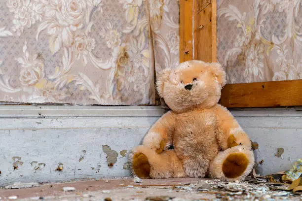 A dirty teddy bear sitting in the corner in an old abandoned house. The wallpaper is peeling, and the floor is littered with debris.