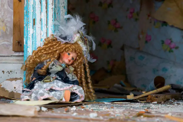 An old doll sittibg against the door frame in an abandoned house. Debris litters the floor, and wallpaper is peeling from the walls in the room behind.
