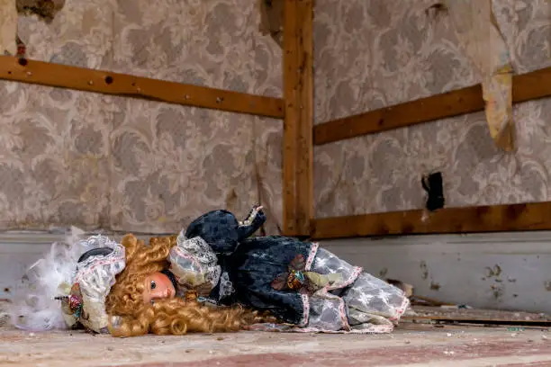 An old doll lying on the floow of an abandoned house. Wallpaper peels from the wall and the floor is dirty.