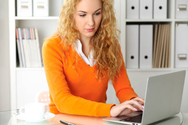 istock Young beautiful business woman or student communicates by laptop computer while sitting at the desk and holding black pen. She Orange sweater is at the girl. Education, business and lifestyle concept 941160670