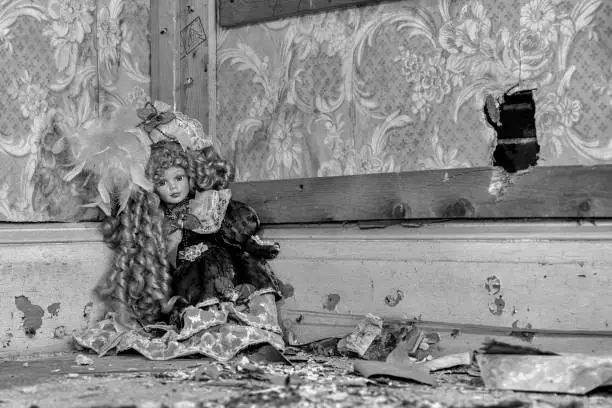 An old dall sitting ina corner in an abandoned house. There is a hole in a wall, and the floor is littered with dirt and debris. Black and white.