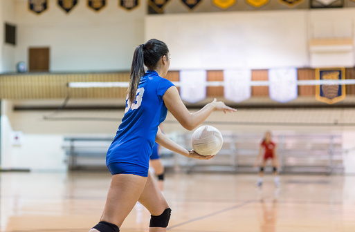 A teenage volleyball player gets ready to serve\\ the ball in a high school gym. The shot is from behind her. The opposing team is getting ready to return the serve. There are banners hanging in the gym overhead.