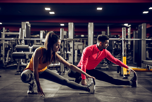 Young healthy sporty active shape girl with a ponytail doing leg stretches on the floor while crouching with a handsome helpful personal trainer next to her in the gym.