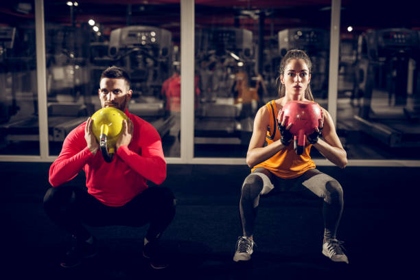 schließen sie nach vorne ansicht konzentriert und motiviert starke junge fitness paares in der sportswear mit kettlebells umgekehrt und kniebeugen in der turnhalle in der nacht zu tun. - coach individuality organized group gym stock-fotos und bilder