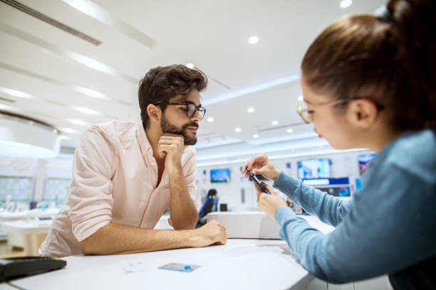 istock Close up view of stylish bearded hipster curious man getting instructions for a mobile from professional girl seller in a tech store. 941212570