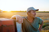 Senior agronomist man standing in a field leaning against old tractor after harvest at sunset.