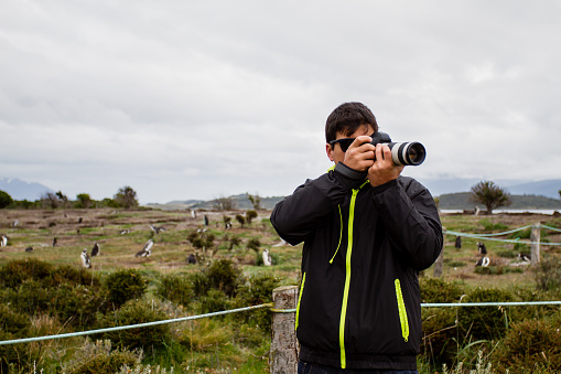 Photographer standing with camera in hand in Martillo Island, with many penguins in the background, Ushuaia, Patagonia.