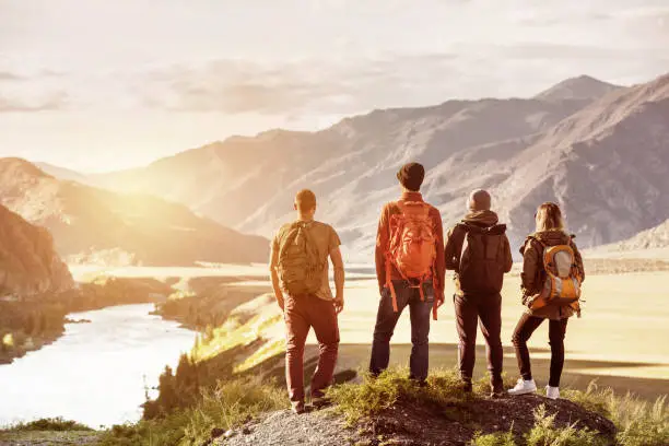 Four friends stands on view point and looking at sunset mountains and river. Adventures travel concept with space for text