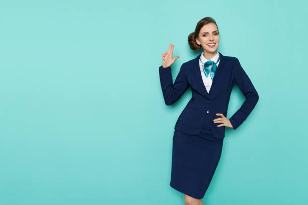 Smiling Stewardess Is Showing Pistol Hand Sign Happy stewardess in blue formal wear is showing pistol hand sign, smiling and looking at camera. Three quarter length studio shot on turquoise background. air stewardess stock pictures, royalty-free photos & images