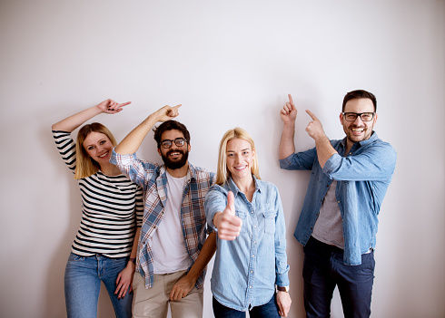 Group of young handsome colleague people standing against the wall showing at empty editable space above while one girl standing with thumb up and looking at the camera.