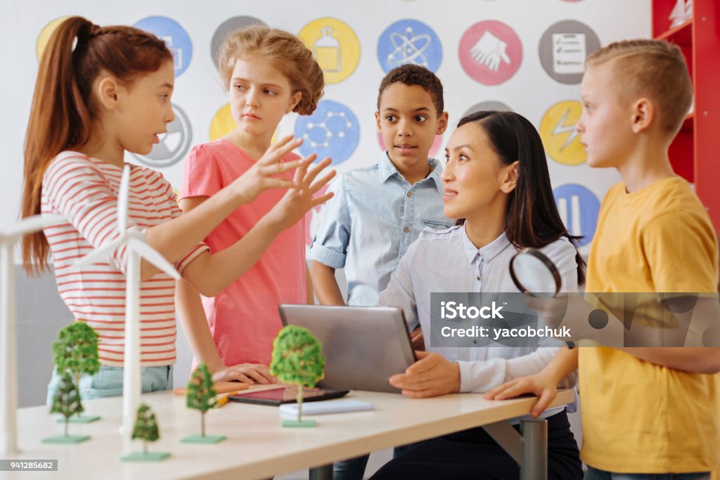 Smart schoolchildren sharing their ecology project idea with teacher Little geniuses. Pleasant smart schoolchildren sharing their ecology project idea with their teacher while she listening attentively to the group leader Child Stock Photo