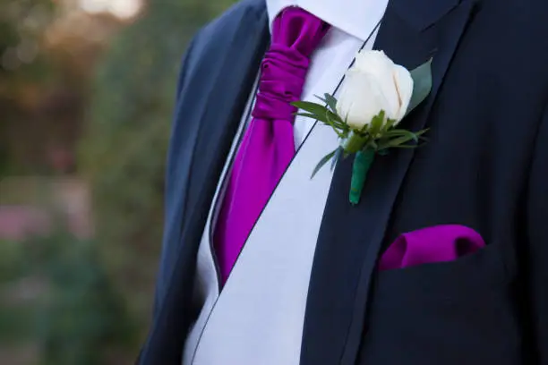 detail of a suit of groom with a white rose in the lapel of the blue suit with gray vest and tie and purple handkerchief