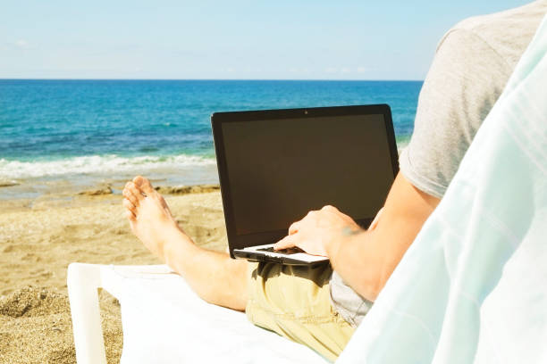 istock Young man in grey shirt sitting at the beach, working on laptop. Blogger, writer, coder, remote worker surfing browsing researching on notebook. Wireless internet on sea shore. Background, copy space 941306636