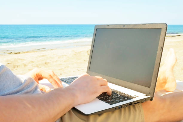 istock Young man in grey shirt sitting at the beach, working on laptop. Blogger, writer, coder, remote worker surfing browsing researching on notebook. Wireless internet on sea shore. Background, copy space 941306666