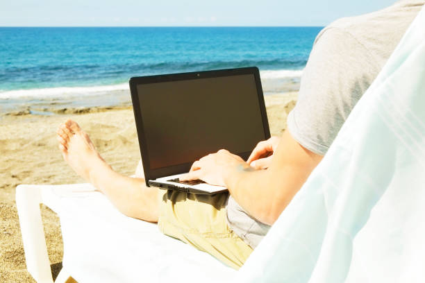 istock Young man in grey shirt sitting at the beach, working on laptop. Blogger, writer, coder, remote worker surfing browsing researching on notebook. Wireless internet on sea shore. Background, copy space 941306668