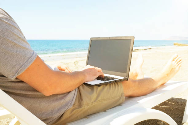 istock Young man in grey shirt sitting at the beach, working on laptop. Blogger, writer, coder, remote worker surfing browsing researching on notebook. Wireless internet on sea shore. Background, copy space 941306706