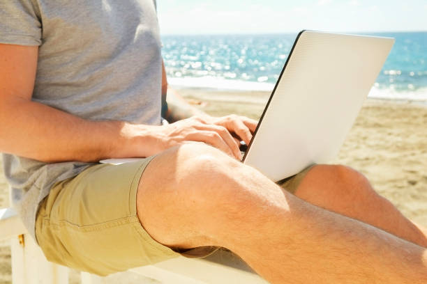 istock Young man in grey shirt sitting at the beach, working on laptop. Blogger, writer, coder, remote worker surfing browsing researching on notebook. Wireless internet on sea shore. Background, copy space 941306818