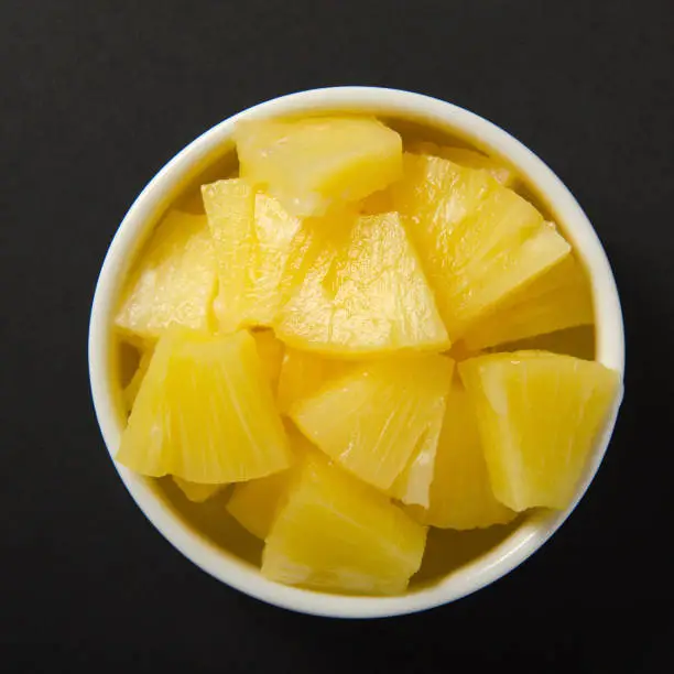 Photo of Pieces of pineapple cut into slices in a white bowl. Ripe yellow sweet fruit on black background