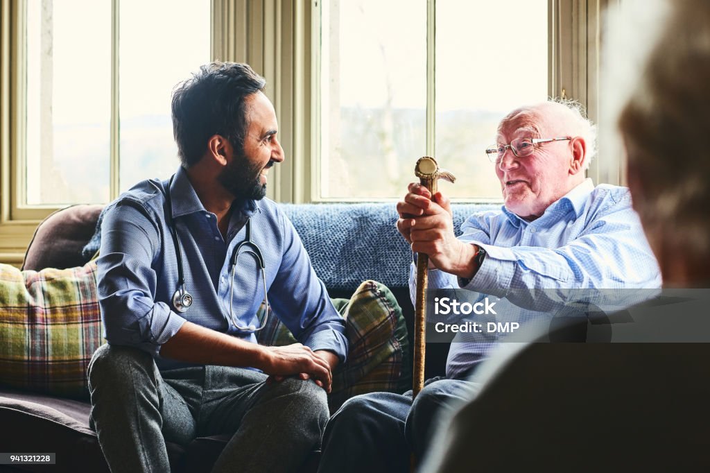 Smiling doctor visiting senior man at home Young male doctor and senior man sitting on sofa and smiling during home visit Senior Adult Stock Photo