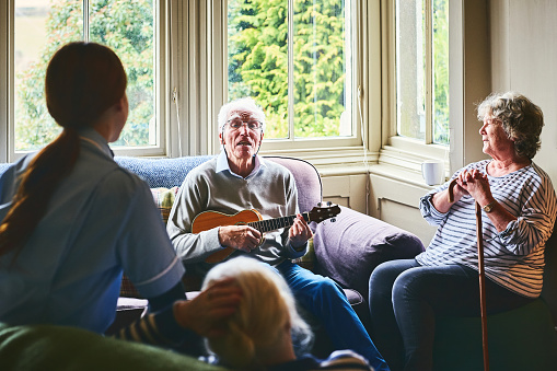Senior people enjoy at nursing home, elderly man playing guitar with a woman and nurse listening.
