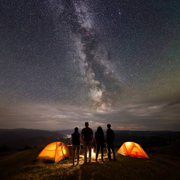 Photo of Rear view two young couples at night in tent camp stand enjoying starry sky, Milky way, mountains and luminous town