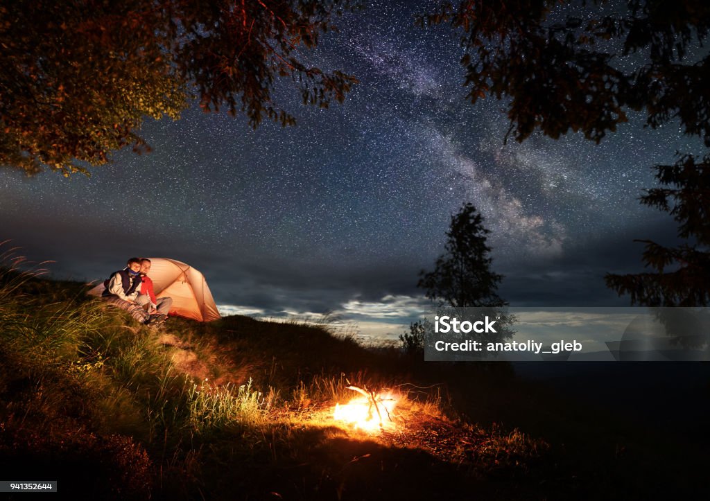 Couple amoureux se trouve sur la colline près de tente appréciant brûle le feu sous le ciel nocturne parsemé d’étoiles brillantes - Photo de Au loin libre de droits